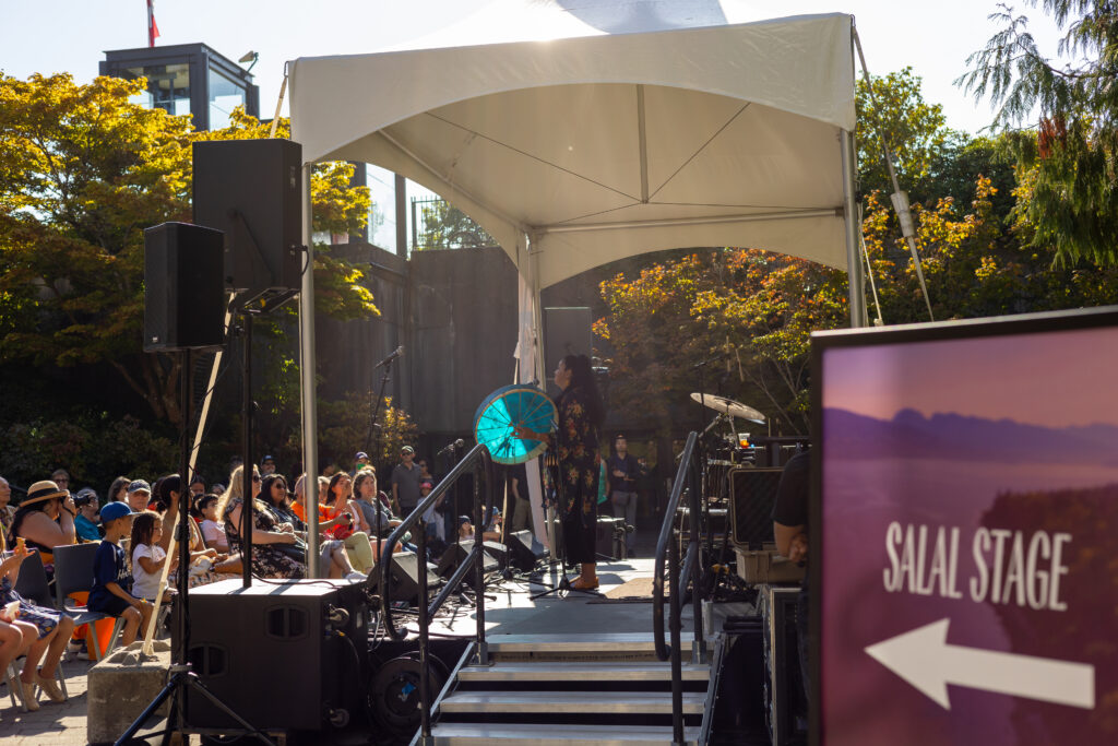 Audience watching a performance at the Salal Stage with the festival sign visible in the foreground.