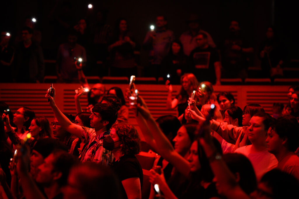 Audience members holding up phones with flashlights lit during an indoor performance at the festival.