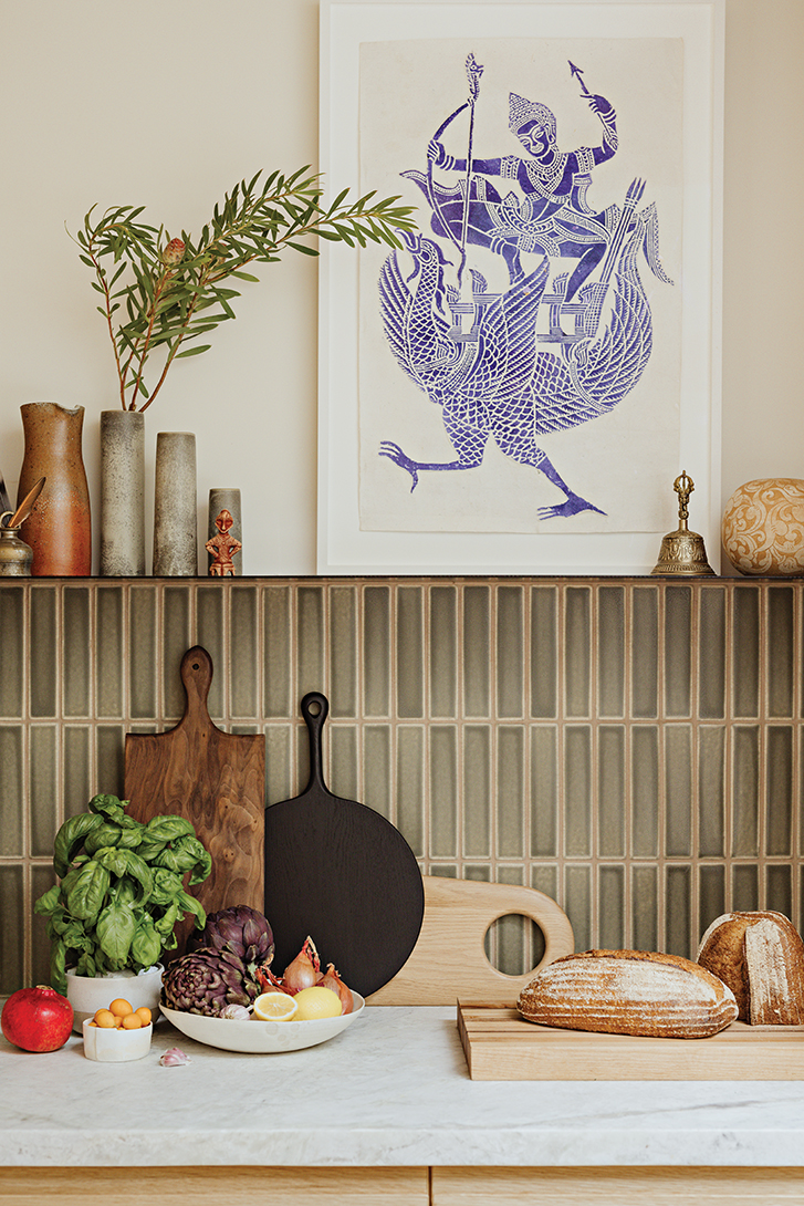 a print of a hindu god riding a chicken is framed on the wall, hung above a row of green tiles and a marble countertop. bread and cutting boards sit on the countertop.