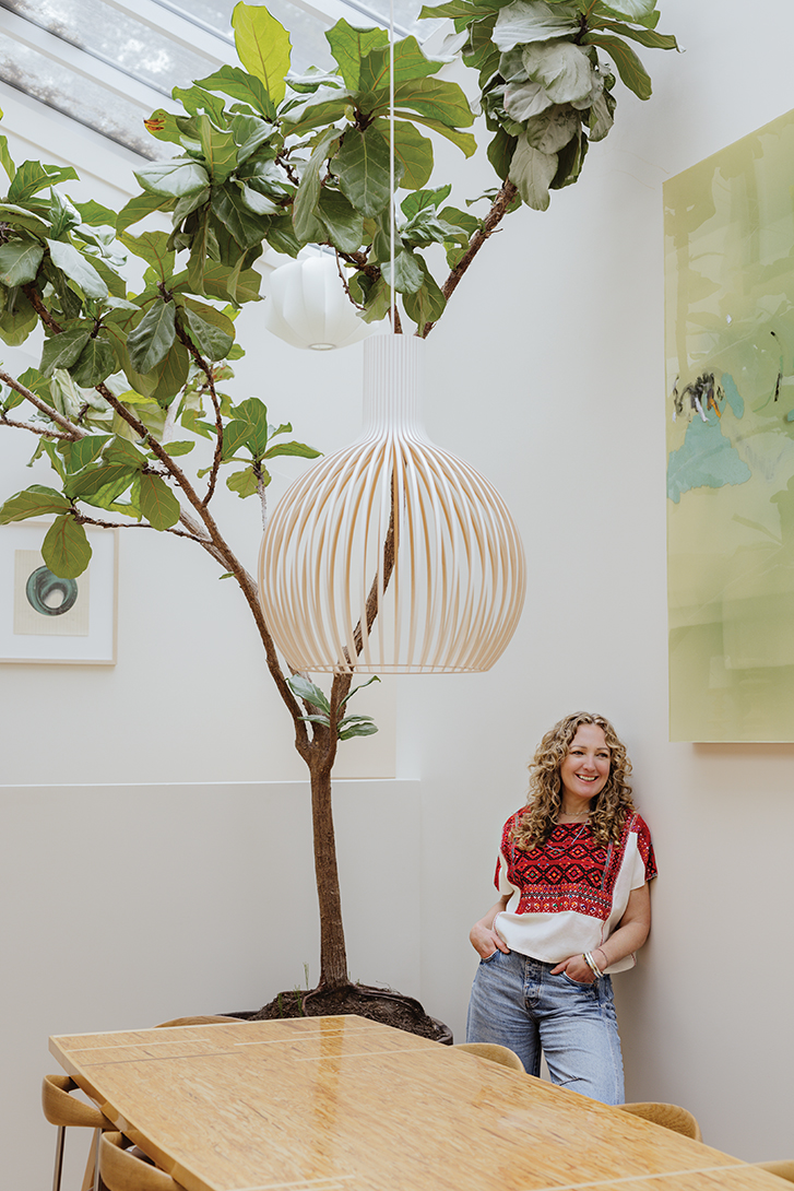 karri stands smiling next to a tall tree growing in a pot inside her dining room. it's very tall and reaches to the skylight.