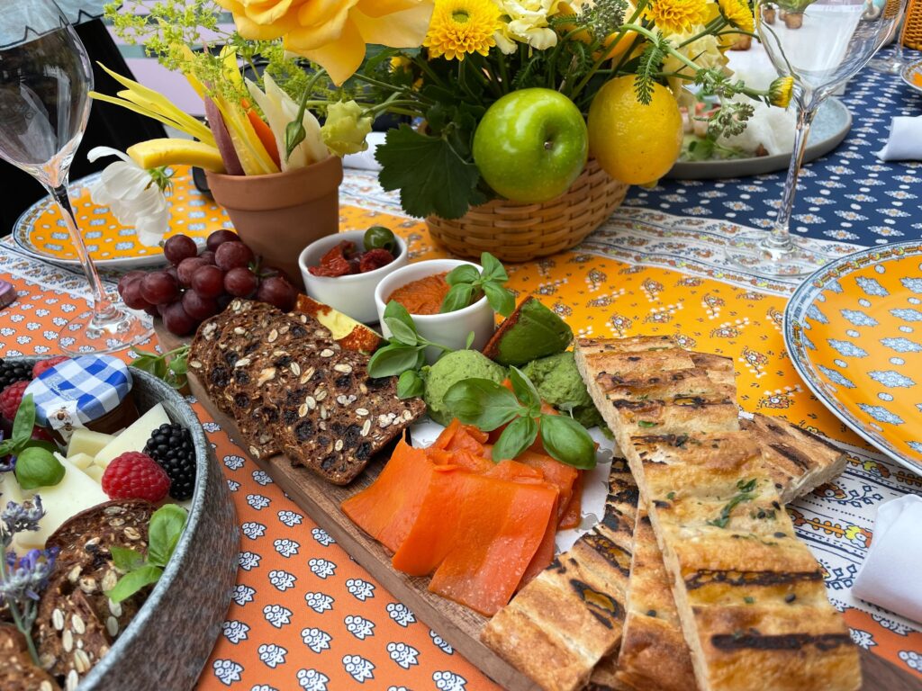 A spread of smoked salmon, grilled flatbread, fruit and crackers, set on a patterned tablecloth with a yellow floral centrepiece
