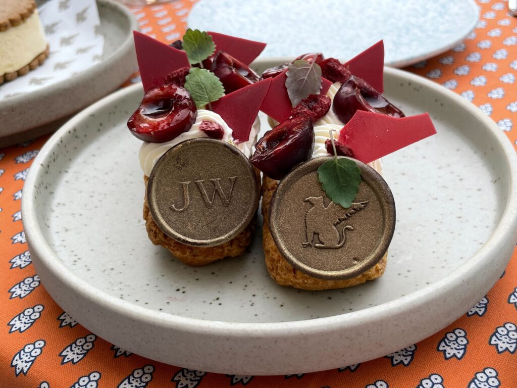 Plated éclairs topped with red chocolate shards, cherries and two JW chocolate coins, served on an orange floral tablecloth.