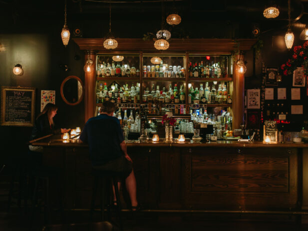 A dimly lit bar with warm lighting, featuring a wide selection of bottles on shelves behind the bar. Two patrons are seated at the bar, engaged in quiet conversation. The bar is adorned with vintage light fixtures and a small chalkboard menu on the wall.