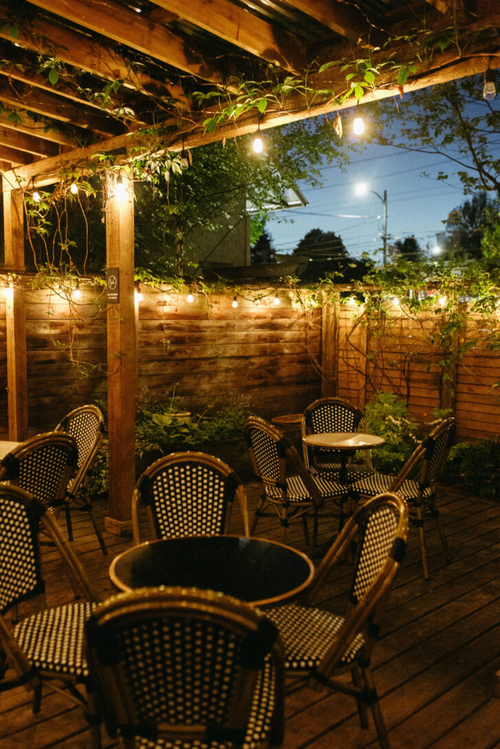 Charming outdoor patio at night, featuring string lights hanging from a wooden pergola adorned with climbing plants. Cozy round tables with wicker chairs are arranged on the wooden deck, creating a warm and inviting atmosphere for evening relaxation.