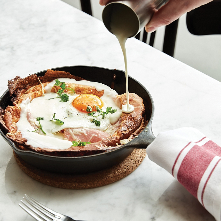 a hand is pouring cream onto a fluffy 'dutch baby' style pancake in a skillet 