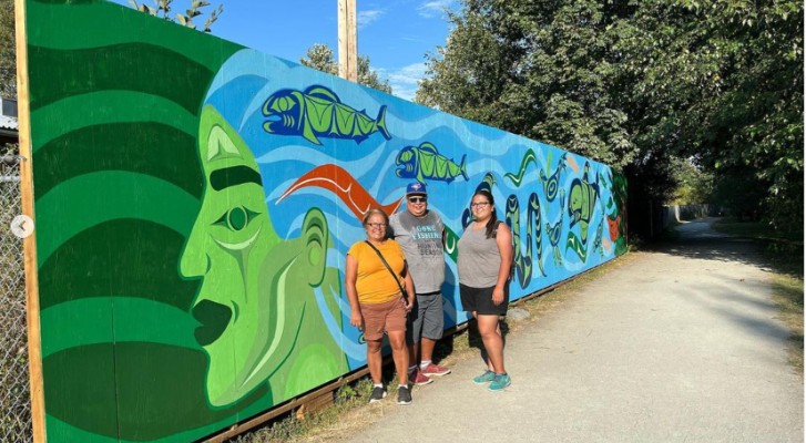Artist Olivia George in front of a mural she painted. The mural is a face in the color green with blue hair that represents the ocean 