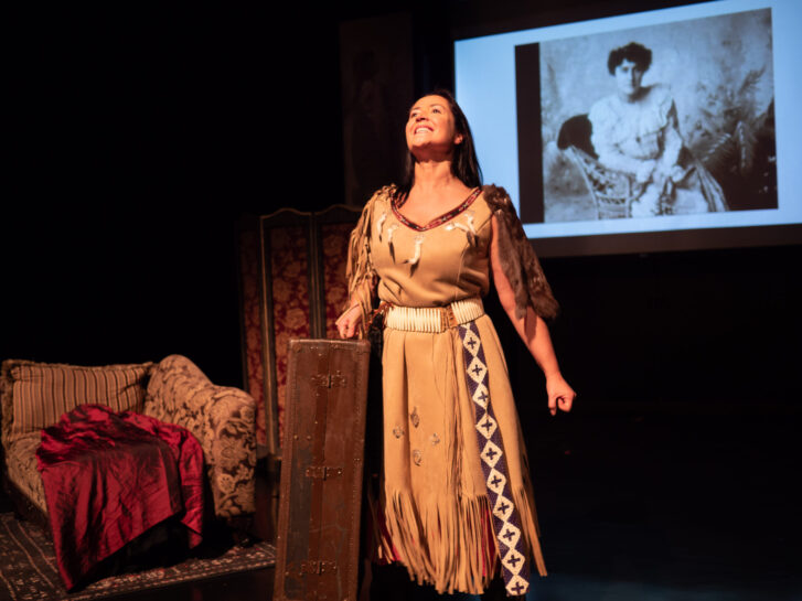 Cheri Maracle, dressed in traditional attire, performs on stage as Pauline Johnson. She stands with a suitcase, looking upwards, with a historic photo of Pauline Johnson projected in the background. The stage is set with a vintage couch and ornate screen.