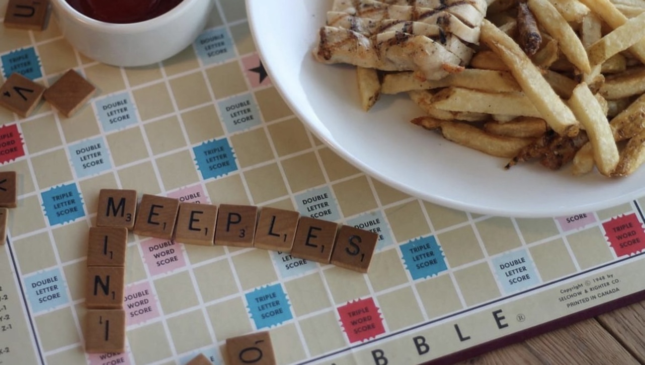 Scrabble tiles spelling "Meeples" next to a plate of chicken and fries.