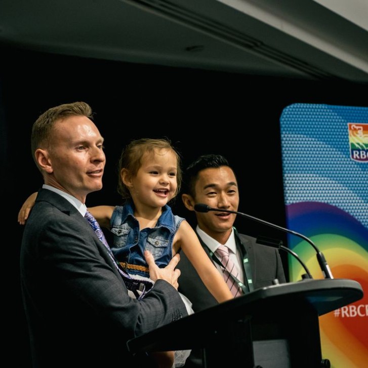 Two men in suits are giving a speech while holding a smiling child. 