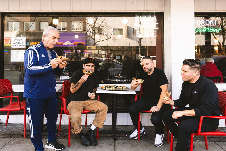 Four men eating pizza outside of the Don't Argue pizza restaurant.