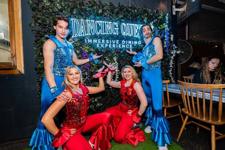 4 performers in costume and bellbottoms posing in front of a neon sign that reads "Dancing Queen Dining Experience" 