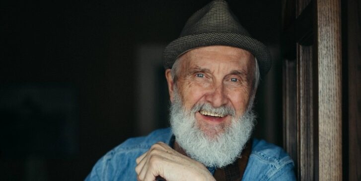 Portrait of an older man (Fred Penner) with a grey beard and a hat, smiling warmly. He is wearing a denim shirt and leaning slightly against a wooden surface.