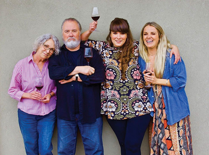 Catherine Coulombe of vinAmité Cellars and family. Four people standing in front of a light grey wall, smiling and holding glasses of red wine. From left to right: a woman with grey hair in a pink checkered shirt, a bearded man in a black shirt with his arms crossed, a woman in a floral-patterned top holding a wine glass above her head, and a woman in a blue cardigan over a colourful dress.